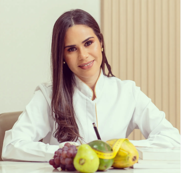 Dra. Carina Malta, nutricionista, sorrindo em jaleco branco com frutas frescas sobre a mesa